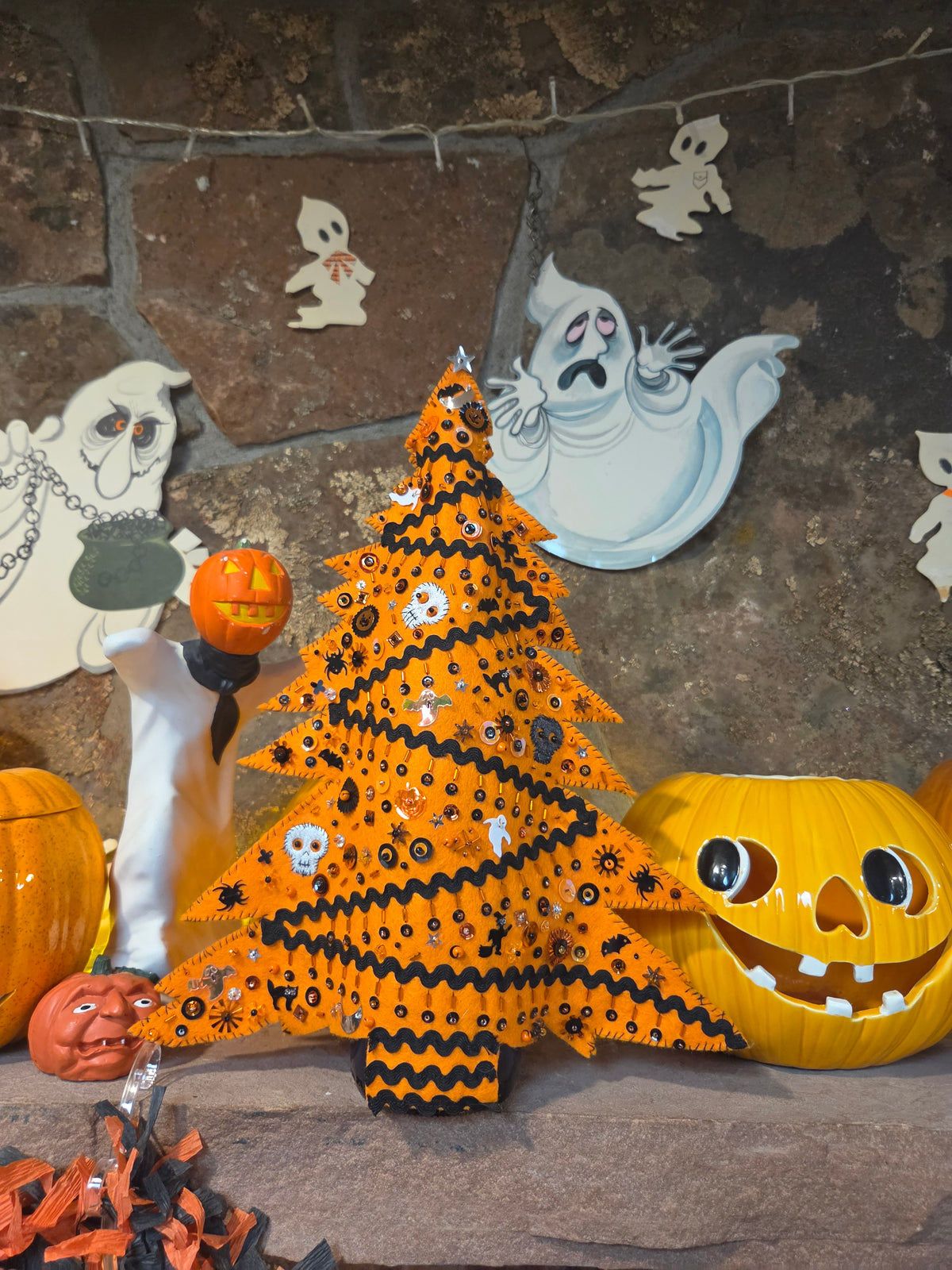 An orange Halloween Christmas tree decorated with black zigzag stripes, white skulls, and small black spider motifs, styled on a stone hearth with pumpkins and ghost decorations.