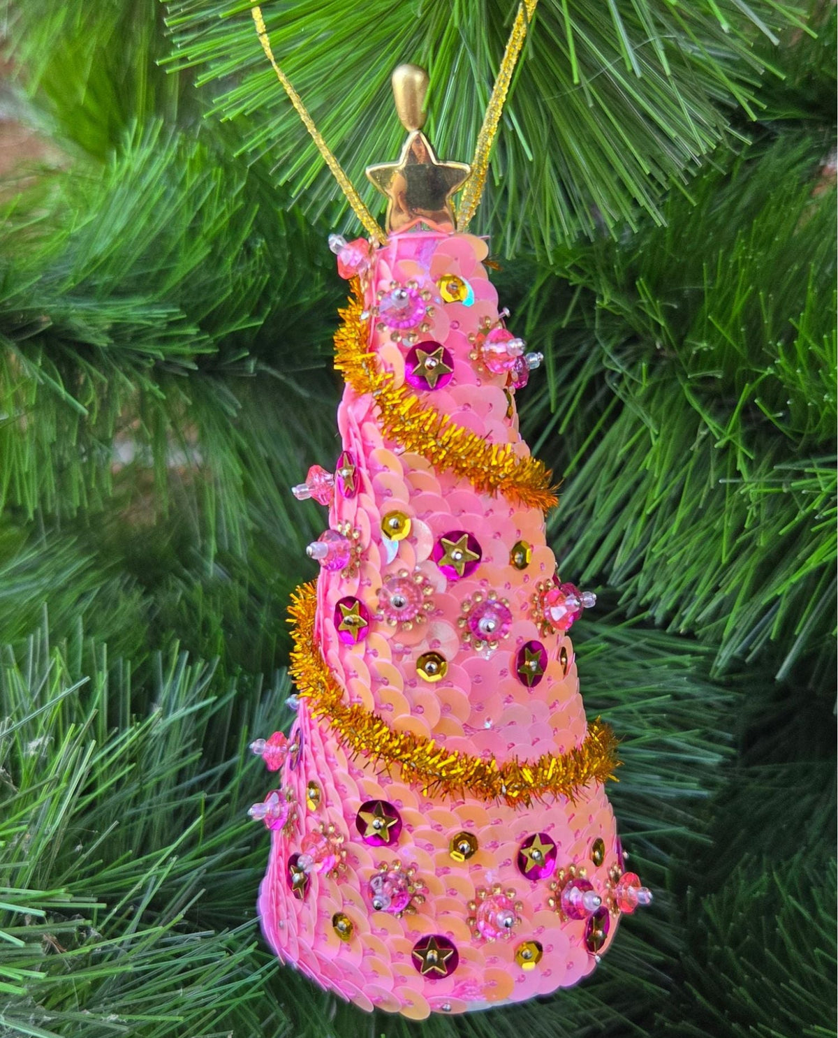 A pink Christmas tree ornament shaped like a tiered dress, decorated with gold tinsel, pink and purple rhinestones, and a gold star topper, hanging on a green Christmas tree branch.